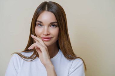 Çekici, koyu renk saçlı kadının yakın plan çekimi, beyaz tişört giymiş, kapalı alanda bej arka planda izole edilmiş pozlar veriyor. İnsanlar, güzellik, sağlık konsepti