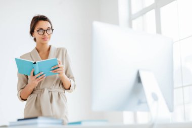 Zarif giyimli genç, koyu renk saçlı bir kadın, mavi not defterini monitöre odaklamış, iş stratejisi üzerine kafa yoruyor, şeffaf gözlük takıyor, geniş beyaz kabinede poz veriyor.