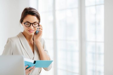 İş kadınının yatay görüntüsü partneriyle çalışma programını modern akıllı telefondan tartışıyor, projeye çözüm buluyor, not defterine odaklanıyor, gözlük takıyor ve resmi takım elbise giyiyor, kapalı alanda duruyor.