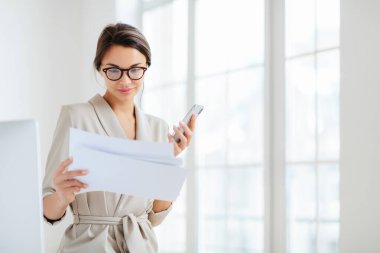 Belgelere odaklanmış, hazırlanmış raporları dikkatle okuyan, modern akıllı telefonları olan, şeffaf gözlükler takan ve ofis içi çeklerin üzerine poz veren kadın çalışanlar.