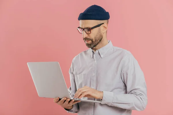 Stock image Indoor shot of busy male freelancer wears optical glasses for good vision, holds modern electronic gadget, surfes internet, develops new website, isolated over pink background. Technology concept