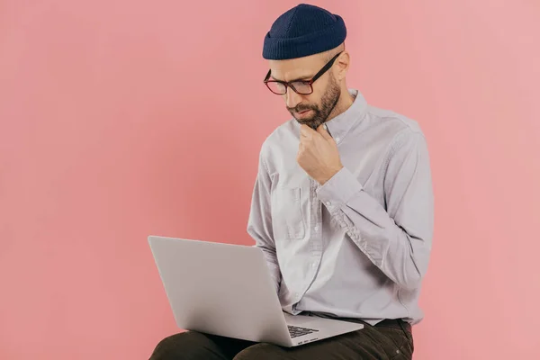 stock image Photo of concentrated unshaven man holds chin, looks attentively at screen of laptop computer, enjoys online communication, wears optical glasses and formal outfit, isolated over pink background