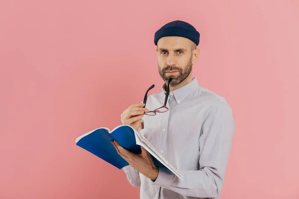 Self confident serious man with dark stubble, takes off spectacles, has attentive look at camera, holds opened textbook, learns necessary information, poses against pink background. Education