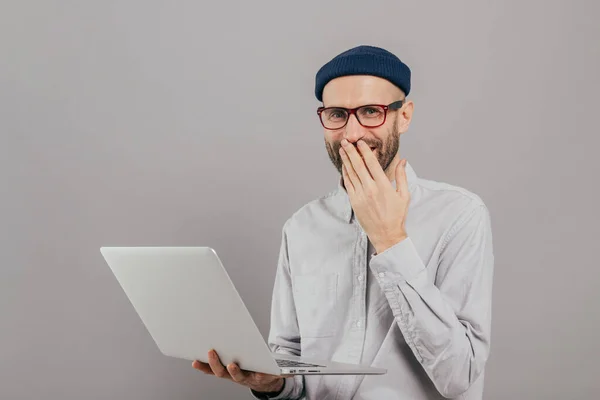 stock image Talented male graphic designer works remotely on gadget, laughs positively, covers mouth with palm, wears spectacles and white shirt, stands against grey background. People and work concept.