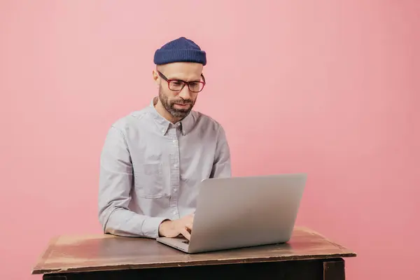 stock image Concentrated hipster student reads information and checks data, focused in monitor of laptop computer, keyboards something, sits at desk, wears formal clothes, isolated over pink studio wall