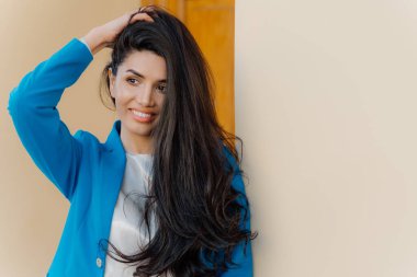 Fotoğraf iyi görünümlü muhteşem esmer kadın başında el tutar, uzun koyu saçlı bir tarafta taranmış, mavi resmi kostüm giymiş, bilgi için boş alan ile beyaz duvar yakınında duruyor