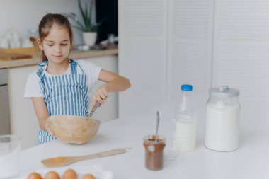 Çocuklar, yemek pişirme ve ev konsepti. Güzel, siyah saçlı bir kız kasede malzeme çırpıyor, pastaya hamur hazırlamakla meşgul, geleceğin aşçısı oluyor, beyaz tişört ve çizgili önlük giyiyor,