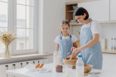 Anne ve kızının birlikte yemek pişirdiği yatay bir çekim. Anne küçük kıza aşçılık dersi veriyor. Lezzetli yemek hazırlamak için un, yumurta ve süt kullanıyor. Aile zamanı, pişirme ve aşçılık konsepti