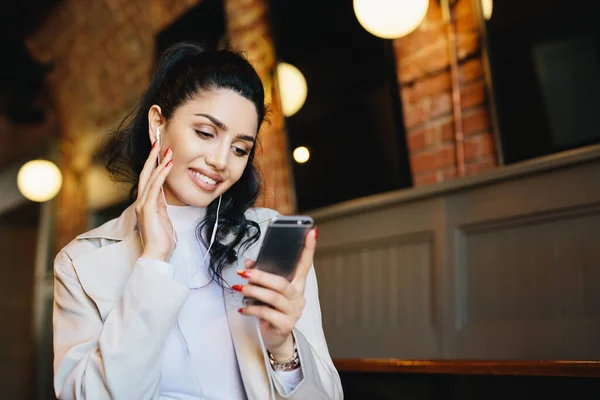 Smiling female with dark hair tied in pony tail wearing white clothes holding smartphone while listening to music on her electronic device. Stylish woman resting in cafe using app having smile on face