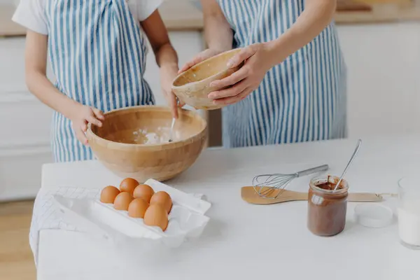 stock image Cropped image of mothers and daughters hands mixing ingredients to prepare dough and bake tasty pastry, stand near kitchen table with eggs, melted chocolate in glass, wear striped blue aprons