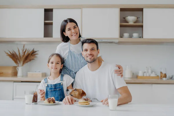 stock image Smiling lovely mother and wife embrces with love daughter and husband, prepared delicious breakfast for them. Little child and father, their pedigree dog eat pancakes in kitchen. Friendly family