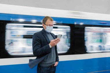 Gözlüklü ve koruyucu tıbbi maskeli bir adamın fotoğrafı, modern telefon kullanıyor, toplu taşıma araçlarıyla seyahat ediyor, platformda hareket halindeki trene karşı duruyor, yeni Covid-19 virüsü hakkında bilgi topluyor.