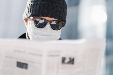 Yetişkin bir adamın güneş gözlüğü, şapka ve steril sağlık maskesi takıp, gazete okurken, dünyadaki durumu öğrenirken, koronavirüs yayılıp, bulanık arka planda poz verirken yakın plan çekimleri.