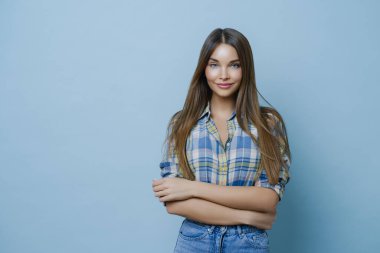Uzun düz saçlı, kareli gömlekli, kot pantolonlu, ellerini çapraz tutan, temiz ciltli, minimum makyajlı, pozitif duygularını ifade eden, fotoğraf çekiminden hoşlanan çekici genç bir kadının yatay görüntüsü.
