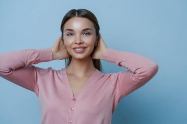 Mükemmel, temiz tenli, dişlek, nazik gülüşlü, ellerini saçından ayırmayan, pembe kazak giyen, stüdyoda fotoğraf çeken, mavi arka planda poz veren güzel bir kadının portresi. Kozmetik ve güzellik kavramı
