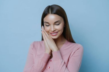 Mutlu, utangaç bir kadının portresi ellerini yüzüne bastırır, konsantre olur, iltifat duymaktan memnun olur, etkileyici bir ifadesi vardır, şefkatli bakışları vardır, mavi arka planda izole edilmiş.