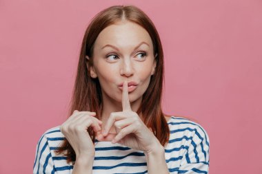 Merak beyaz bir kadın sessizlik işareti yapar, işaret parmağını ağzına tutar, gizlice bir yana görünüyor, çizgili rahat kıyafetler giyer, metniniz için boş alan ile pembe studio duvarın üzerinden teşkil etmektedir. Sessiz ol