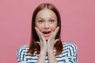 Olumlu mutlu sürpriz beyaz bir kadın bir şey inanılmaz görür, yanaklarda ellerini tutar, hayretle, rahat kıyafetler, pembe bir arka plan karşı modelleri giyinmiş ağız açılır. Reaksiyon kavramı