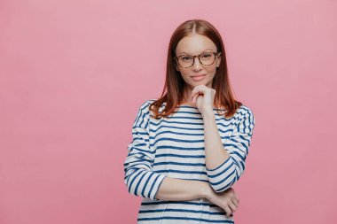 Çekici koyu saçlı kadın kapalı atış eller çene altında tutar, çizgili denizci kazak, optik gözlük takıyor, pembe bir arka plan üzerinde modeller, doğrudan kameraya benziyor, kendine güvenen ifadesi olan