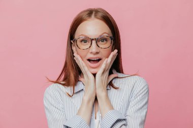 İnsan yüz ifadeleri, reaksiyon kavramı. Güzel başarılı iş kadını yanaklarda iki elini tutar, zarif beyaz çizgili gömlek giyer, yüz ifadesi, pembe duvardan izole şaşırttı