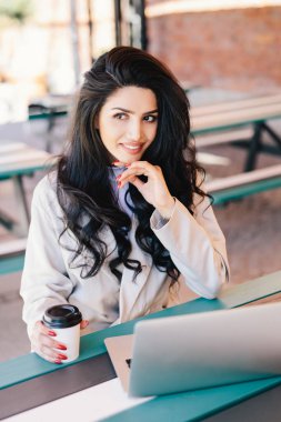 Karanlık gözlere sahip uzun dalgalı saçlı beyaz genç kadın serbest meslek ve gizemli bir şekilde kenara uzaktan çalışma modern dizüstü pc, ücretsiz wi-fi lezzetli kahve içme kullanarak sağlıklı cilt arıyorsunuz.