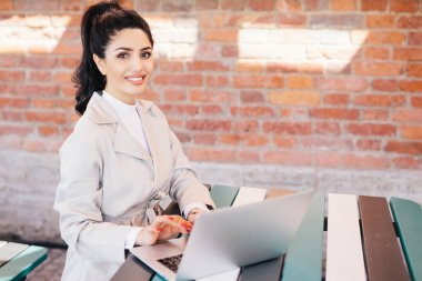 Oldukça esmer öğrenci kadın gülümseyerek kameraya bakarak onun tez yazma bilgisayarda çalışan zarif Beyaz önlük giyiyor. İşkadını kapalı tuğla duvar oturan zarif giysiler