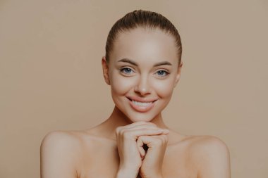 Günlük makyajlı, dişleriyle gülümseyen ve ellerini çenesinin altında tutan, yarı çıplak bir şekilde içeride dikilen, estetik ameliyatlardan sonra kendini yenilenmiş hisseden yetişkin bir kadının yakın çekimi. Güzellik ve yüz bakımı kavramı