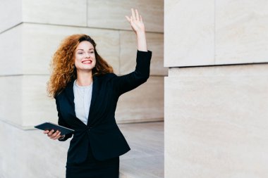Gülümseyen mutlu iş kadını resmi giyinmiş, elinde modern tablet bilgisayarla elini sallayarak arkadaşını fark ediyor, onu gördüğüne seviniyor. Kıvırcık saçlı kadın 
