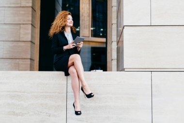 Portre siyah takım elbise ve modern tablet ile oturan ince uzun bacakları olan yüksek topuklu ayakkabılar içinde zarif güzel kadın bir kenara mutlu bir ifadeyle bakıyor. Açık poz işçisi kadın ofis