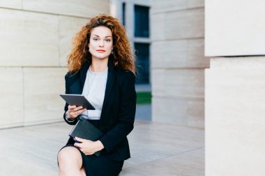 Zarif Bayan Beyaz bluz, siyah ceket ve etek giymiş office builduing yanında otururken tablet ve cep kitabı tutarak portresi. Kıvırcık saçlı, güzel yüzünü başarılı iş kadını