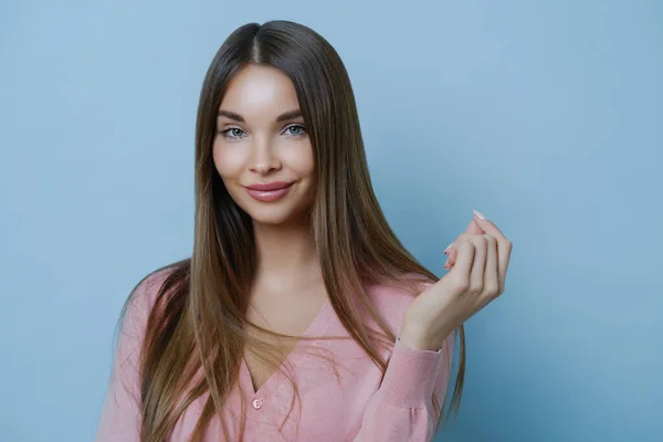 Beautiful young European lady with no blemishes and makeup, has cute look, keeps hand raised, feels delighted, poses against blue background, wears rosy jumper, going to participate in fashion show