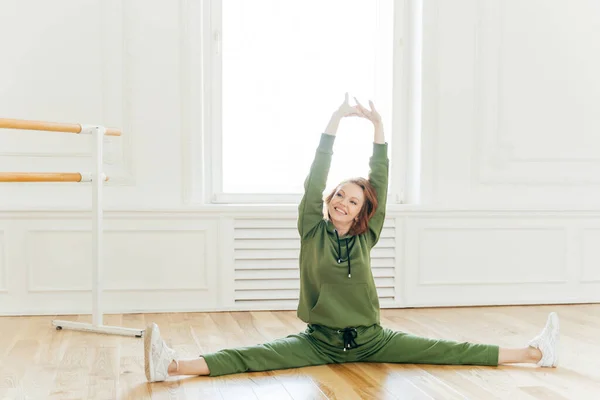 stock image People, strength, fitness, gymnastics concept. Glad sportswoman with cheerful face, raises hands, spreads legs and does splits, stretches in studio. Young dancer has work out, dressed in tracksuit