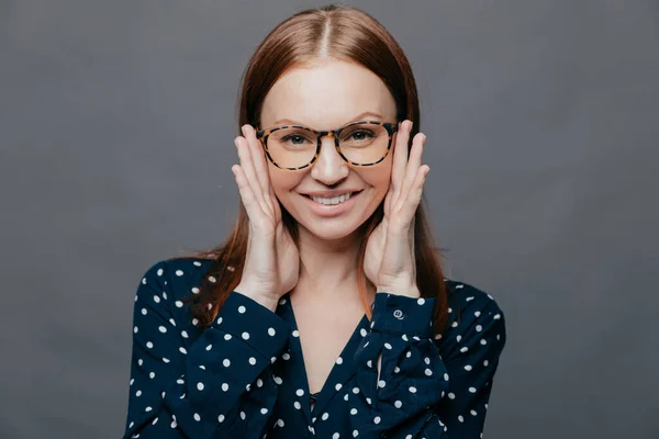 stock image Photo of attractive woman with pleased facial expression, keeps both hands near cheeks, wears polka dot shirt, poses over grey studio wall, being happy. European pleased lady expresses happiness