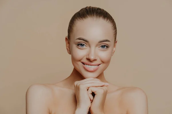 stock image Close up shot of adult woman with fresh daily makeup, smiles toothily and keeps hands under chin, stands half naked indoor, feels refreshed after cosmetic procedures. Beauty and face care concept