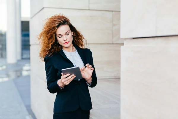 stock image Elegant lady with curly hair, wearing black suit, typing messages or making business report while using tablet computer. Female entrepreneur working on new business project. Business and career