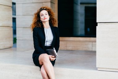 Çok güzel kadın modeli gür dalgalı saçlı, kırmızı dudaklar ve parlayan gözleri, resmen tablet cep kitabı, güvenle kameraya bakarak ile tutarak bacaklarını geçerken giyinmiş. İş kavramı