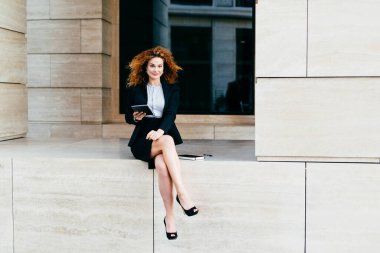 İş, kariyer ve başarı kavramı. Zarif ince genç işkadını siyah takım ve oturan, yüksek topuklu ayakkabılar bacak modern gadget, tutarken bacak güvenle kameraya bakıyor