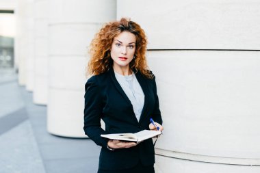 Yatay siyah takım elbise ve beyaz bluz, elinde kalemiyle defter tutan giyen ciddi güzel işkadını kıvırcık saçlı, ince kaşlar ve kıvırcık saçlı, portresi doğrudan kameraya bakıyor