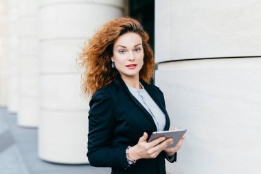 Siyah takım elbise ve beyaz bluz giyen, tablet bilgisayar holding, mesajlar yazarak veya açık ayakta iken Internet arama kıvırcık saçlı güzel kadın. Modern cihaz ile güzel iş kadını