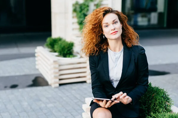 Pretty businesswoman with curly hair, pure skin and red lips, wearing formal costume while sitting at outdoor cafe or terrace, touching her modern table, communicating with friends using free wi-fi