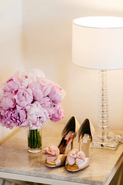 漂亮的鞋子 高跟鞋 灯和有站在床头桌上的粉红色花朵的花束 花束和新娘的鞋子 婚礼鞋和鲜花 特殊的场合的做准备 — 图库照片