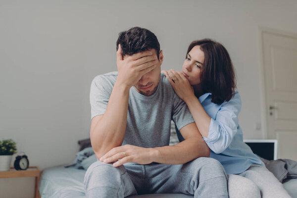 Attractive female consoles her sad boyfriend who has depression and some problems, pose at bedroom on bed, sit against domestic interior. Wife and husband try solve difficult situation in life.
