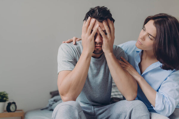 Stressful young man cries from despair, loving mother tries to calm him, pose together at bed against domestic interior, have some problems. Upset husband sees no way out in difficult situation