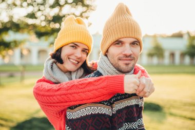 Şapka ve sıcak pamuk kazaklı genç erkek geri duran kocası kucaklayan memnun gülümsüyor, hafta sonları birlikte geçirmekten keyif, Bahçe veya park yürüyüşler, splendid güneşli sonbahar hava hayranım