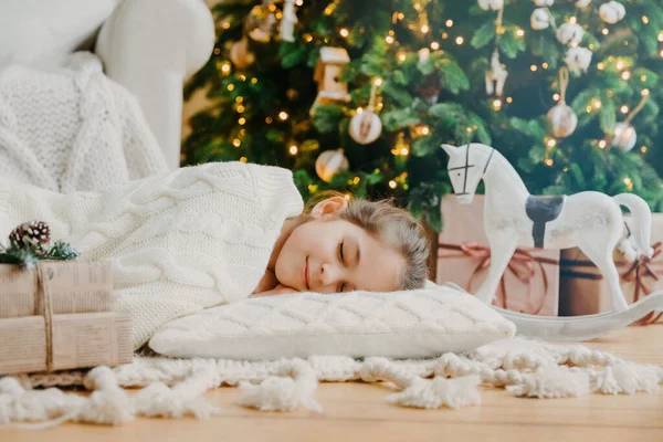 Çekici kız süslemeli yeni yıl ağacına karşı yerde yumuşak beyaz yastıkta uyuyor. Oyuncak at ve hediye kutularıyla çevrili güzel rüyalar görüyor. Çocuklar, dinlenme ve kış tatili konsepti.