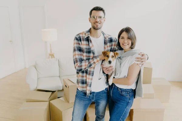 Affectionate family couple celebrate moving day, embrace and holds favourite pet, enjoy relocaton and starting living together, pose in big room among carton boxes. Mortgage and relocation concept