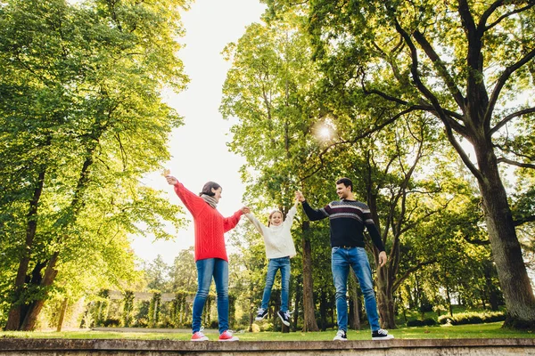 stock image Portrait of friendly affectionate family have fun together, spend free time outdoors, stand against green trees in park, try to rock small child in hands. Three family memebers play on nature