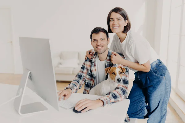 图片上 快乐的黑发女微笑着靠着丈夫的肩膀 他们在电脑上键盘 在网上搜索新公寓 与家犬在宽敞的房间里摆姿势 — 图库照片