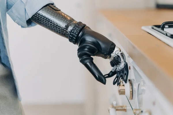 Stock image Disabled person's electronic cyber hand switches on stove in kitchen. Amputee with high-tech carbon arm, finger and wrist joint sensors. Daily routine of disabled individual.