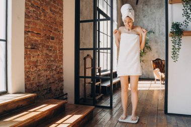 Kadın evde elektronik teraziyle ağırlık ölçer. Zayıflatıcı hedef başarısını kutlar, sevinçle el kaldırır. Başarılı kilo kaybı.